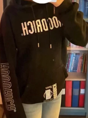 Svart hoodie från Hoodrich - Säljer en svart hoodie från Hoodrich med stor vit logga framtill och mindre tryck på fickan. Klassisk huva med snören och magficka. Materialet känns mjukt och bekvämt, perfekt för en avslappnad streetwear-look.