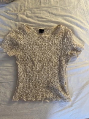 Krämvit smockad topp Gina - Säljer en krämvit smockad topp från Gina Tricot med korta ärmar och rund halsringning. Toppen har en stretchig struktur och vågiga kanter vid ärmslut och nederkant. Perfekt för en trendig och avslappnad look.