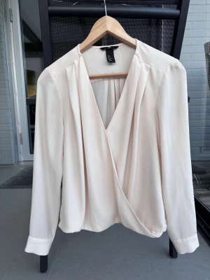 Krämvit omlottblus från H&M - Säljer en stilren krämvit omlottblus från H&M med djup v-ringning och långa ärmar. Blusen har lätt drapering framtill och breda manschetter vid ärmsluten. Perfekt att matcha med jeans eller kjol för en trendig look.