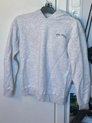 Grå hoodie med tryck Ma chérie - Ljusgrå hoodie med huva och trycket 'Ma chérie' på bröstet och ett större hjärta med pil och text på ryggen. Mjuk och skön, perfekt för chill dagar. Ribbstickade muddar vid ärmslut och nederkant.