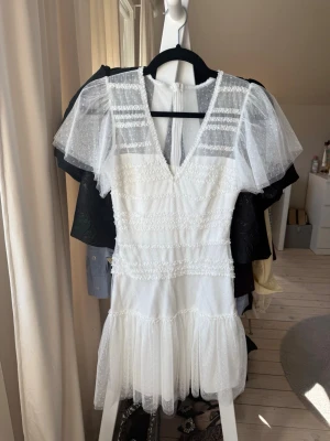 Vit kort klänning med volang och spets - Superfin vit kort klänning med v-ringning, volang nertill och transparenta kortärmade partier i mesh med prickigt mönster. Klänningen har dragkedja bak och detaljer i spets. Perfekt för student och sommarens alla tillfällen från bubbleroom. Väldigt fint skick och använd endast en gång. Storlek xss.