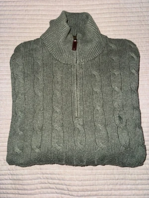 Grå kabelstickad tröja Polo Ralph Lauren - Kabelstickad grå tröja från Polo Ralph Lauren med halv dragkedja och hög krage. Tröjan har långa ärmar, ribbstickade muddar och diskret broderad logga på bröstet. Perfekt för lager-på-lager och snygg till jeans.