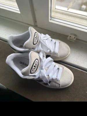 Vita Adidas sneakers med svarta ränder - Snygga vita Adidas sneakers med klassiska svarta ränder på sidorna och stor Adidas-logga på plösen. Skorna har vita snören och ovandel i mockaliknande material. Perfekta för en sportig och trendig look.