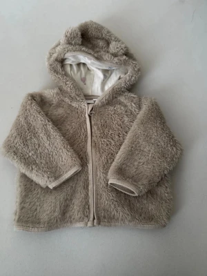 Beige teddyjacka med huva - Mysig teddyjacka från Newbie i beige färg med huva och dragkedja. Jackan har ett fluffigt material och ett innerfoder med ballongmönster. Perfekt för att hålla sig varm under kyliga dagar.