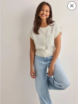 Vit kortärmad kofta med knappar - En stilren vit kofta med korta ärmar och knappar framtill. Koftan har en mjuk och lite fluffig struktur, ribbade kanter och en rak passform. Perfekt att matcha med jeans för en avslappnad look.