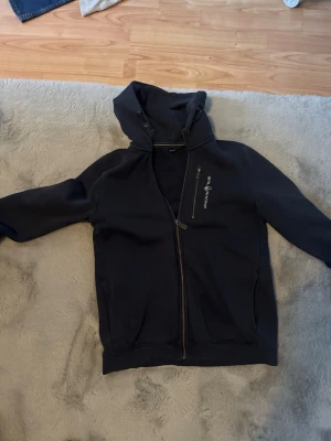 Marinblå hoodie från Sail Racing - Snygg marinblå hoodie från Sail Racing med dragkedja i guld och broderad logga på bröstet. Hoodien har huva och en snedställd dragkedja framtill samt en ficka med dragkedja. Perfekt för dig som gillar sportig och stilren design.