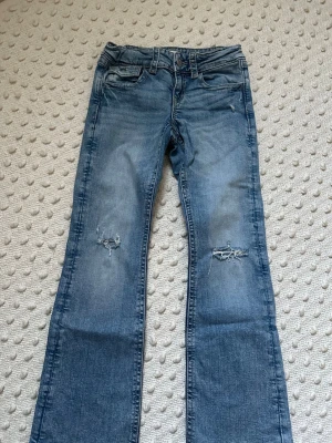 Blå bootcut jeans med slitningar - Säljer ett par blå bootcut jeans med slitningar på knäna och snygg tvätt. Jeansen har klassiska fem fickor, bakfickor med lock och knapp samt låg midja. Perfekta för en avslappnad och trendig look.