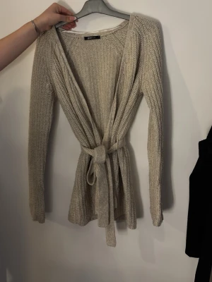 Beige stickad kofta med knytskärp - Säljer en beige stickad kofta från Gina tricot  med långärmad design och knytskärp i midjan. Koftan har ett öppet framstycke och är gjord i ett mjukt, grovt stickat material som passar perfekt för lager-på-lager. Enkel och stilren look.