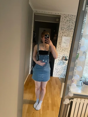 Blå jeansklänning med hängslen - Cool kort jeansklänning med hängslen och fickor framtill. Klänningen har en klassisk blå denimfärg och är ärmlös med justerbara axelband. 