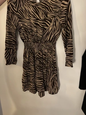 Kort zebra-klänning med resår - Säljer en kort klänning med zebramönster i beige och brun. Klänningen har långärmade ärmar och resår i midjan som ger en snygg passform. Perfekt för dig som gillar djurmönster och vill sticka ut. Materialet känns mjukt och lätt.