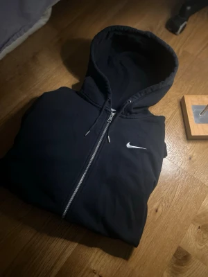 Svart Nike zip-hoodie med huva - Säljer en svart hoodie från Nike med dragkedja och huva. Hoodien har den klassiska Nike-loggan broderad i vitt på bröstet och snören i huvan. Perfekt för dig som gillar sportig och enkel stil.