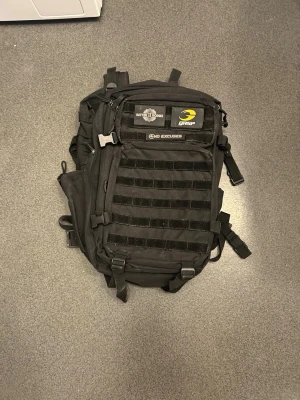 Svart ryggsäck från GASP - Ryggsäck i svart slitstarkt material från GASP med flera fack, justerbara remmar och MOLLE-panel på framsidan. Utrustad med patchar och texten 'NO EXCUSES'. Perfekt för gym, sport eller outdoor-aktiviteter.