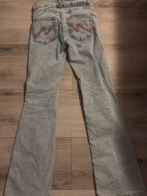 Ljusblå slitna bootcut jeans - Coola ljusblå jeans med bootcut passform och slitningar framtill. Bakfickorna har röda zigzag-broderier som ger extra edge. Perfekta för dig som gillar en avslappnad och trendig look. Köptes för 699, storlek EU 34💓 använda få gånger 