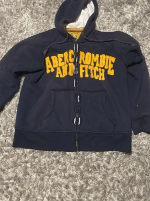 Marinblå hoodie från Abercrombie - Säljer en marinblå hoodie från Abercrombie & Fitch med stora gula bokstäver på bröstet. Hoodien har dragkedja, huva med snören och en stor magficka. Materialet är mjukt och insidan är ljus. Perfekt för dig som gillar klassisk college-stil. Har ett hål där back 