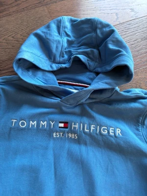 Blå hoodie från Tommy Hilfiger - Snygg blå hoodie från Tommy Hilfiger med broderad logga och text 'EST. 1985' på bröstet. Klassisk huva och ribbade detaljer. Materialet är mjukt och bekvämt, perfekt för en avslappnad look. Ikoniska Hilfiger-färger syns på insidan av halsen.