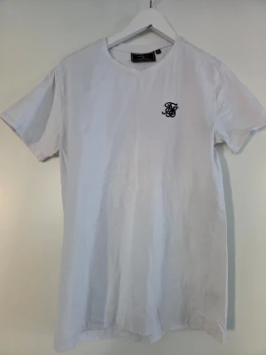 Vit t-shirt från SikSilk - Klassisk vit t-shirt från SikSilk med rund hals och korta ärmar. T-shirten har en stilren svart logga broderad på bröstet och är tillverkad i mjuk bomull för skön komfort. Perfekt basplagg till garderoben.