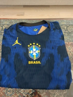 Blå Brasilien matchtröja Jordan - Snygg helt ny blå Brasilien matchtröja från Jordan med gula detaljer och CBF-logga på bröstet. Tröjan har ett unikt mönster i blått och svart, rund halsringning och gula ränder längs axlarna. Perfekt för fotboll eller träning. DMA FÖR MER INFO (viktig info, pris kan diskuteras och bilder kan skickas privat.)