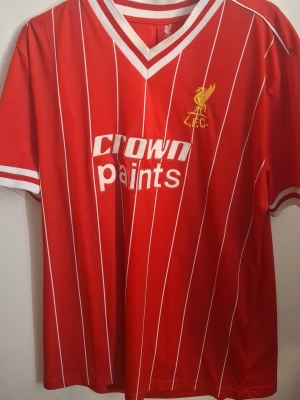 Liverpool retro matchtröja röd XL - Äldre Klassisk röd Liverpool FC matchtröja med vita tunna ränder och vit V-ringad krage. Tryck med 'Crown Paints' på bröstet och klubbens logga i gult. Korta ärmar med vita detaljer. Perfekt för dig som älskar fotboll och retrostil.
