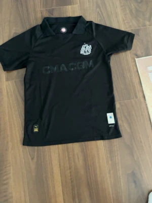 Svart fotbollströja från Puma - Säljer en svart fotbollströja från Puma med Marseille-logga på bröstet och CMACGM-tryck på framsidan. Tröjan har korta ärmar, krage och är tillverkad i ett lätt och funktionellt material som passar perfekt för träning eller match.