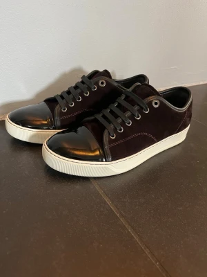 Svarta sneakers med lackdetalj - Snygga sneakers i vinröd mocka med glansig lackad tå och vita sulor. Skorna har svarta skosnören och metallfärgade öljetter. Perfekta för dig som vill ha en stilren men ändå unik look. Det är märket lanvin och är i perfekt ny skick. Ny pris kostar skorna 5200. Skon är stor i storleken. Uk6