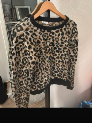 Leopardmönstrad stickad tröja - Snygg stickad tröja med leopardmönster i beige, svart och brunt. Tröjan har svarta ribbstickade muddar vid hals, ärmslut och nederkant. Perfekt för dig som vill sticka ut med ett djurmönster i garderoben.