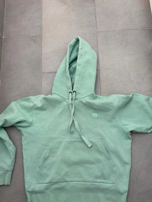 Mintgrön hoodie från Acne Studios - Snygg mintgrön hoodie från Acne Studios med klassisk känguruficka och dragsko i huvan. Tillverkad i mjuk bomull och har Acne Studios ikoniska ansiktslogga på bröstet. Perfekt för en chill och trendig look.
