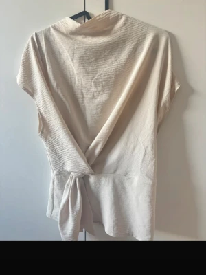 Beige omlottopp med knytband - Snygg beige omlottopp med knytband i midjan och lätt struktur i tyget. Toppen är ärmlös och har en liten knapp i nacken för stängning. Perfekt till jeans eller kjol för en clean och stilren look.