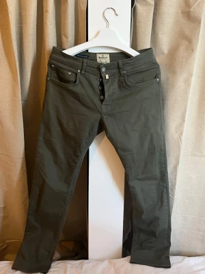 Olivgröna jeans från Morris - Säljer ett par olivgröna jeans från Morris med rak passform och knappgylf. Jeansen har klassisk femficksdesign och diskret detalj med liten tygflik vid fickan. Materialet är mjukt och stretchigt, vilket gör dem riktigt sköna att bära. Perfekta för dig som gillar stilrena och tidlösa jeans.