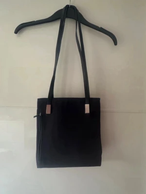 Marinblått fyrkantig axelväska - Stilren  axelväska i fyrkantig modell med långa axelband och silverfärgade metalldetaljer. Väskan har en dragkedjeficka på sidan och är tillverkad i ett slätt tygmaterial. Perfekt för dig som gillar minimalistisk design. Storlek 24x25cm