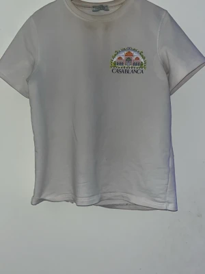 Vit t-shirt från Casablanca - Vit t-shirt från Casablanca med färgglad tryckt grafik på både bröstet och ryggen. Motivet visar en byggnad med palmer och texten 'CASABLANCA'. T-shirten har rund hals och korta ärmar, perfekt för dig som gillar stilrena plagg med coola prints.