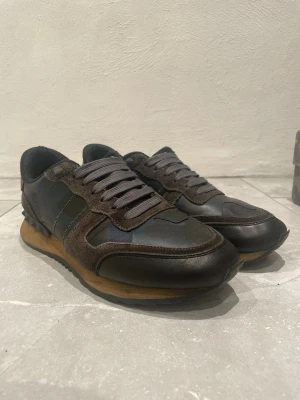 Valentino skor - Säljer ett par snygga svarta sneakers från Valentino Garavani med detaljer i mörkbrunt och grått. Skorna har en låg profil, klassiska snörningar och en kombination av läder och mocka. Yttersulan är beige och boxen med kamouflagemönster ingår. Finns lite defekter, storleken är 40.