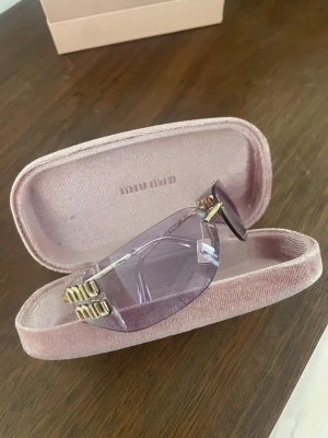 Lila solglasögon från Miu Miu - Snygga solglasögon från Miu Miu med lila tonade glas och guldfärgade detaljer. Modellen har en futuristisk, rektangulär form och transparent båge. Kommer med original rosa sammetsetui och tygpåse med Miu Miu-logga.