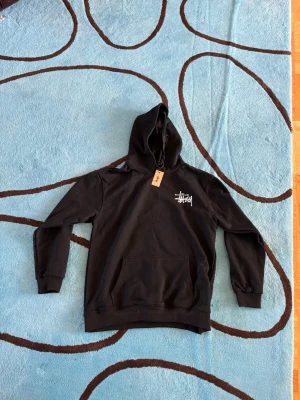 Svart Stüssy hoodie med tärningsmotiv - Cool svart hoodie från Stüssy med vit logga på bröstet och stort tärningsmotiv på ryggen. Klassisk huva med dragsko och stor magficka framtill. Perfekt för dig som gillar streetwear och vill sticka ut med en unik design.