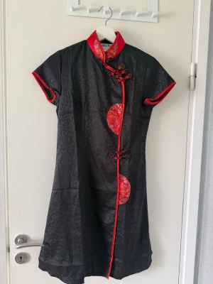 Kinesisk klänning - Snygg svart cheongsam-klänning med röda kanter och dekorativa knappar. Klänningen har korta ärmar och traditionella kinesiska mönster i rött och guld på detaljerna. Ståkrage och figurnära passform ger en elegant look.