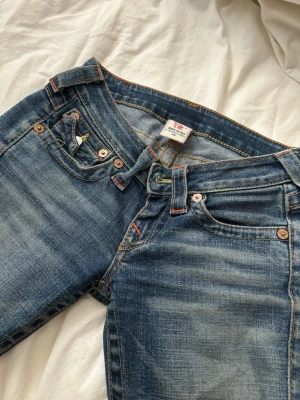 True Religion blå jeans med kontrastsömmar - Säljer ett par blå jeans från True Religion med snygga kontrastsömmar och ikoniska bakfickor med lock och vita knappar. Jeansen har klassisk femficksdesign och slitningar som ger en cool look. Perfekt för dig som gillar streetstyle.