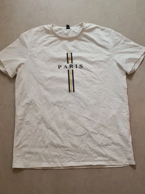 Vit t-shirt med PARIS-tryck - Vit t-shirt med trycket 'PARIS' och två vertikala ränder i svart och beige på bröstet. Klassisk passform och rund halsringning. Perfekt för en clean och stilren look.