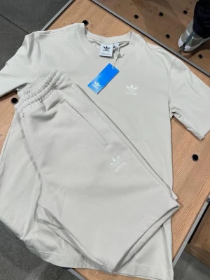 Beige set från Adidas med t-shirt och shorts - Snyggt set från Adidas i beige med matchande t-shirt och shorts. Båda plaggen har Adidas logga framtill och är tillverkade i mjukt material för en avslappnad look. Perfekt för en sportig och trendig stil.