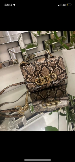 Axelväska i ormmönstrat med guldkedja - Snygg axelväska i ormmönstrat konstläder med guldfärgade kedjedetaljer och justerbar axelrem. Väskan har en rektangulär form och ett stilrent spänne framtill. Perfekt för dig som vill sticka ut med en trendig accessoar.