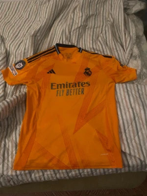 Real Madrid Bellingham matchtröja orange - Snygg orange Real Madrid matchtröja från Adidas med svarta detaljer och trycket 'Emirates Fly Better' på bröstet. Tröjan har Bellingham och nummer 5 på ryggen samt klubbmärke och sponsorloggor. Tillverkad i lätt och ventilerande material, perfekt för fotboll.