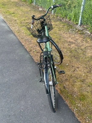Grön damcykel med korg och pakethållare - Snygg grön damcykel med svart sadel, svarta handtag och en praktisk korg framtill. Cykeln har pakethållare bak, stänkskärmar och stöd. Perfekt för att ta sig runt i stan eller till skolan. Metallram och klassisk design.