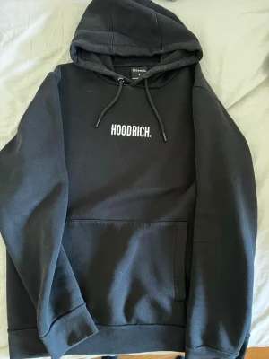 Svart hoodie från Hoodrich - Svart hoodie från Hoodrich med vit logga tryckt på bröstet. Klassisk modell med huva och dragsnören samt stor magficka framtill. Perfekt för en avslappnad streetwear-look. Mjuk bomullsblandning som känns skön mot huden.