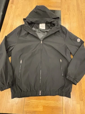 Moncler windbreaker  - Svart vindjacka från Moncler med huva, hel dragkedja och två framfickor med dragkedja. Jackan har elastiska muddar och nederkant samt Moncler-logga på vänster ärm. Tillverkad i lätt, vattenavvisande material som passar perfekt för blåsiga dagar.