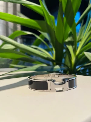Svart Hermé$ armband - Stilrent armband från Hermè$ i silverfärgad metall med svart emalj och ikoniskt H-spänne framtill. Hör av er vid frågor!