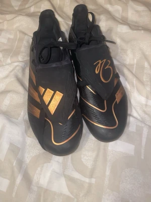 Adidas Predator fotbollsskor svart/guld - Snygga Adidas Predator fotbollsskor i svart med guldiga detaljer och klassiska tre ränder på sidan. Skorna har snörning och en täckt plös med ett guldigt M3-tryck. Tillverkade i syntetmaterial för bra grepp och kontroll på planen.