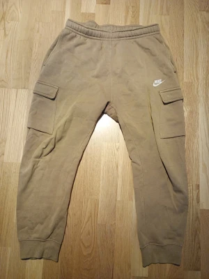 Beige cargo mjukisbyxor från Nike - Beiga mjukisbyxor från Nike med cargofickor på sidorna och broderad vit Nike-logga. Byxorna har resår i midjan och vid bensluten för en skön passform. Perfekta för en avslappnad och trendig stil.