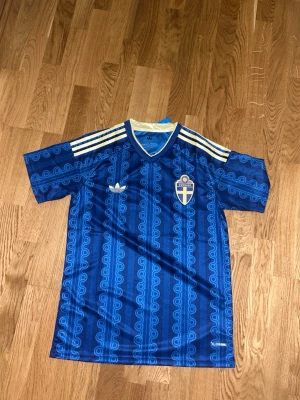 Blå Sverige retro fotbollströja Adidas - Retroinspirerad blå fotbollströja från Adidas med Sveriges landslagsemblem på bröstet. Tröjan har vita Adidas-ränder på axlarna, mönstrad design och V-ringning med vit och blå kant. Perfekt för dig som gillar klassisk sportstil.