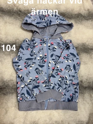 Blommig hoodie med dragkedja - Snygg ljusblå hoodie med blommigt mönster i vitt, rosa och mörkblått. Tröjan har huva, dragkedja framtill och ribbade muddar vid ärmslut och nederkant. Perfekt för dig som gillar färg och mönster🌸 Svaga fläckar vid ärmen. Storlek 104 