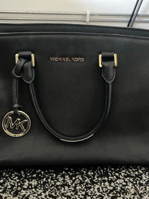 Svart handväska från Michael Kors - Snygg svart handväska från Michael Kors i slätt läder med guldfärgade metalldetaljer och ett MK-hänge. Väskan har två handtag och klassisk, stilren form. Perfekt för dig som gillar tidlös design och vill ha en väska som funkar till allt.