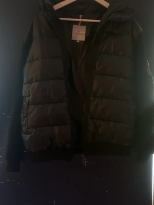 Svart cardigan från Moncler - Snygg svart dunjacka från Moncler med klassisk puffer-design och huva. Jackan har dragkedja framtill, Moncler-logga på ärmen och en ficka med dragkedja. Perfekt för kalla vinterdagar och riktigt stilren look.