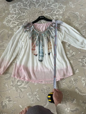 Vit och rosa bohemisk blus med mönster - Luftig blus i vitt och ljusrosa med långa ärmar och bohemiskt mönster runt halsringningen i flera färger. Blusen har en lös passform och är tillverkad i ett tunt, transparent tyg som ger en lätt och somrig känsla.